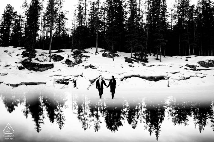 Engaged couple walk at the frozen lake looking at each other an holding hands. Behind them forest which is reflected on the bottom.