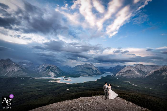 Bride and groom standing next to each other at the top of the mountain. In the background a blue lake and huge Canadian Rockies.