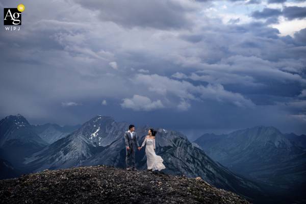 Newlyweds hold their hands on the top on the mountain in Banff. Behind them giant mountain and heavy, storm clouds.