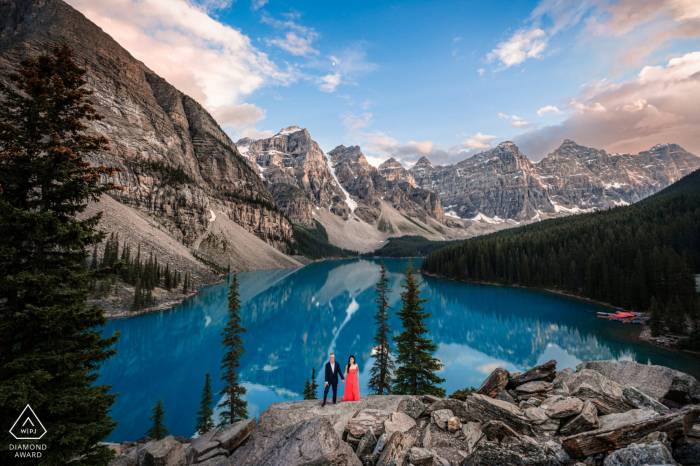 Couple in love holds their hands at the edge on turquoise Moraine Lake in Banff. Behind them huge mountains during the sunset.
