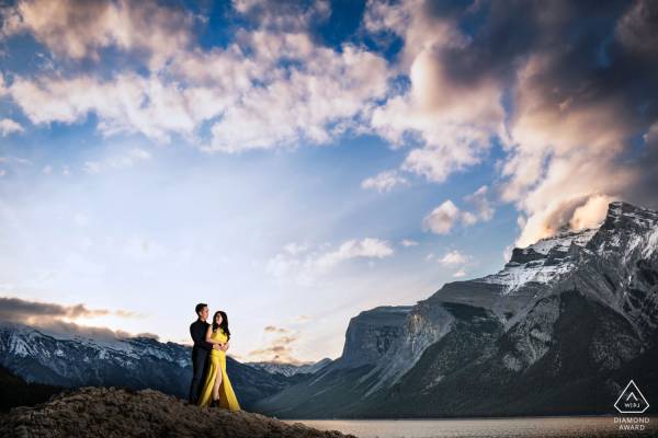 A man hug his fiancé in a yellow dress during stunning sunrise an Minnewanka Lake in Banff.