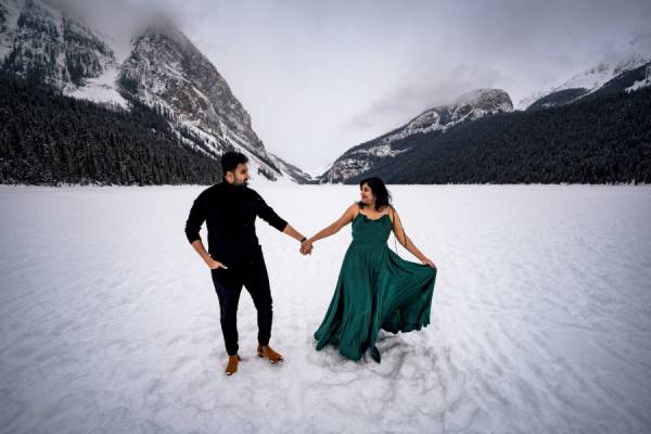 Couple in love holds their hands. Lady wears a green long dress,, men is dressed in black Engagement session at frozen Lake Louise. They are surrounded by Canadian Rocky Mountains.