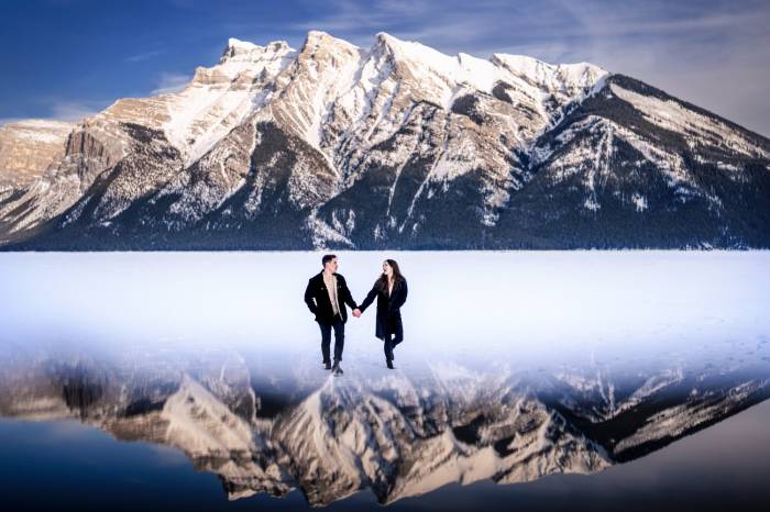 Engaged couple walking on the frozen Minnewanka Lake in Banff holding their hands. On the background huge mountain which is reflected on the bottom. Amazing photograph taken by Calgary Wedding Photographer - 4Eyes Photography.