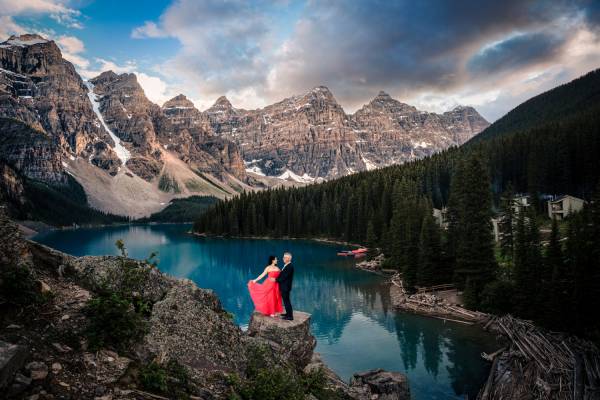 Woman in a red dress and her partner standing on the huge rock. On the background huge Canadian Rockies Mountains and blue Moraine Lake. Photograph taken by Banff Wedding Photographer during the sunset.