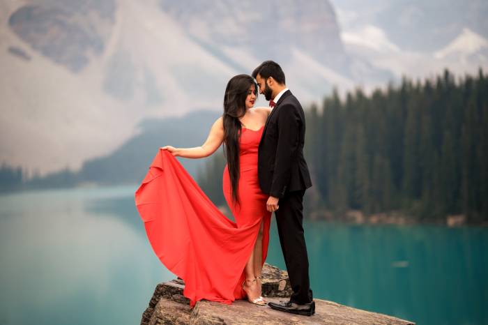 Moraine Lake Engagement Photography. Indian engaged couple standing on the rock. Lady is wearing a red, long dress and her fiancée is wearing a suite.