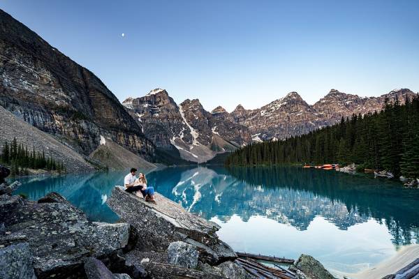 Amazing engagement session at blue Moraine Lake. Couple sits at the rock against the Canadian Rockies and glacier lake.