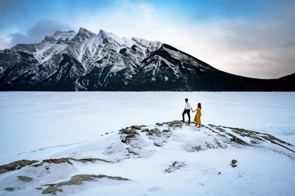 Incredible engagement photography taken by Calgary Wedding Photographer at Minnewanka Lake in wintertime. Couple standing at the giant rock and admire the beauty of the mountains.