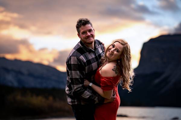 engaged couple look at the camera and smile during sunrise at Minnewanka Lake in Banff