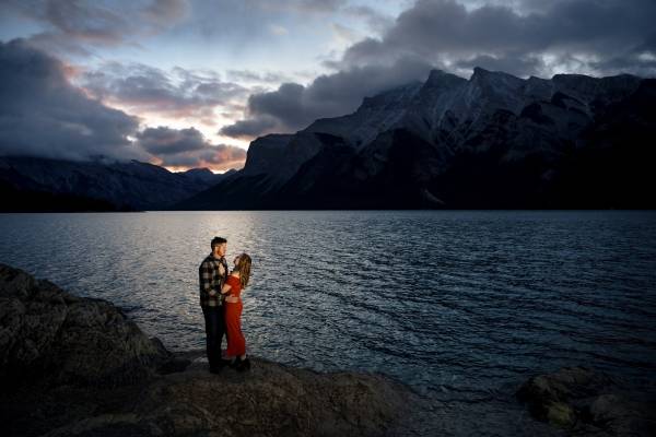 Lady in a red dress and her fiancee look at each other and hug, They are surrounded by the Canadian Rockies at Minnewanka Lake in Banff.