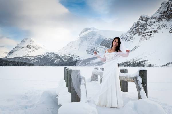 bride standing at the wooden bridge at Bow Lake in Banff. Behind her are tremendous mountains and frozen lake