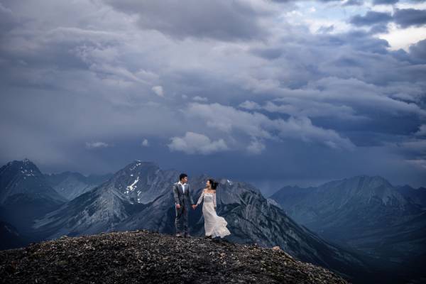 Dramatic Banff Wedding Photograph of the newlyweds holding their hands on the top of the mountain. Being them heave clouds and incredible view of the Canadian Rockies.