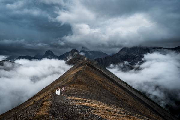 Fearless couple at the top of the mountain walking at the ridge in Banff. On the background stunning Rocky Mountains and heavy clouds.