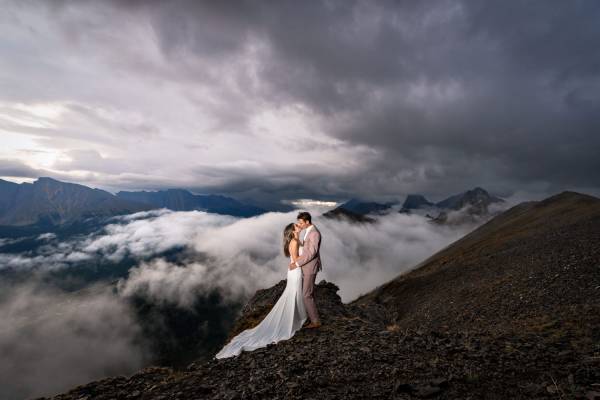 Newlyweds share a kiss on the top of the mountain in Banff during rainy day. Clouds are above and behind them.