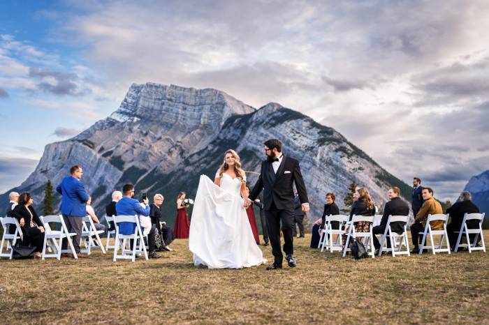 Bride and groom walk holding hands after the ceremony in Banff. On the background guests sitting on the white chairs and huge mountain.