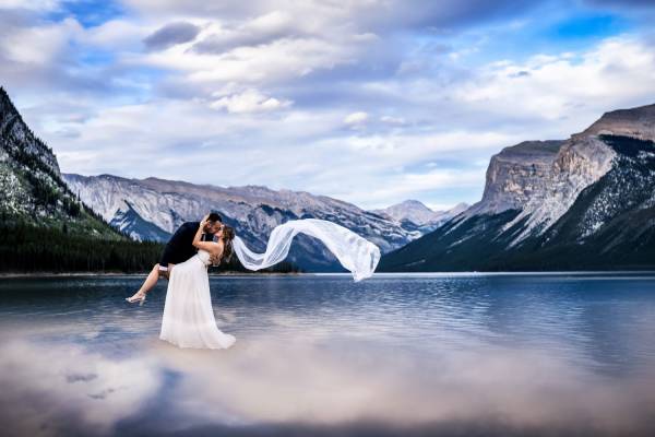 Creative photograph of newly weds dancing at the lake. Bride's veil flow with the air. They are surrounded by the Canadian Rockies at blue Minnewanka Lake . Photo taken by the best wedding photographer 4Eyes Photography.