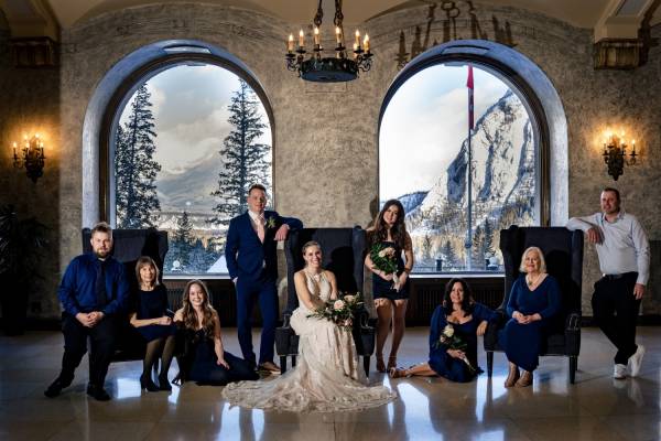 Wedding family portrait in luxury Fairmont Banff Springs hotel. Par of the family sits on the chairs, some of them stand . Behind them huge windows with mountain views.