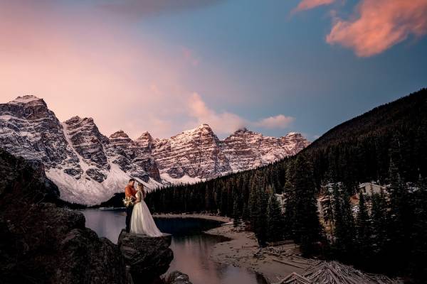 Newlyweds stand at the giant rock at Moraine Lake. They are surrounded by huge Canadian Rockies, Photo was taken during beautiful sunrise by Calgary Wedding Photographer.