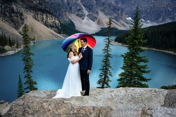 Newlyweds under colourful umbrella. They are posing to Banff Wedding Photographer at Moraine Lake.