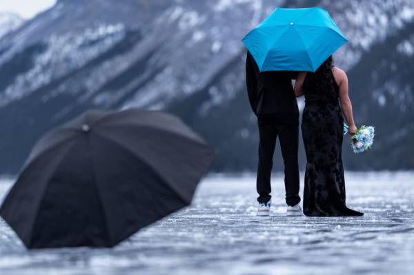 Newly engaged couple walks under a blue umbrella. They look at the beautiful mountain in Banff.