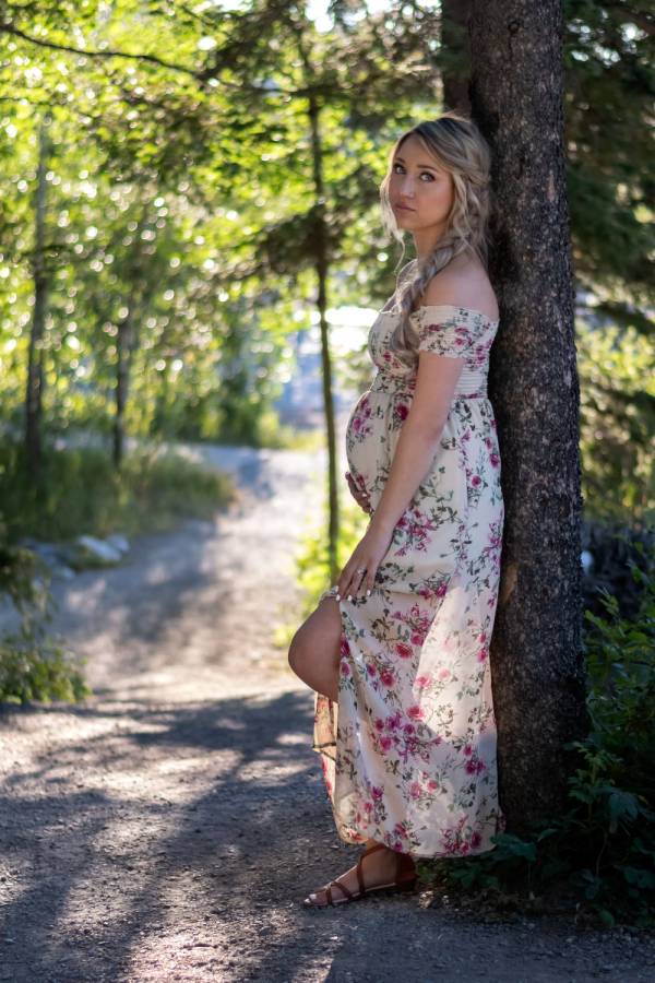 Pregnant woman posing during materning photo shoot in Banff. She lean against the tree and touch her belly.