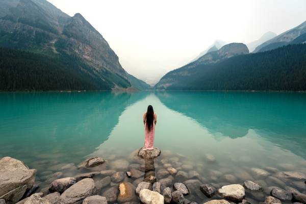 Lady in a pink dress standing at the rock at Lake Louise. She is looking at stunning Canadian Rockies.