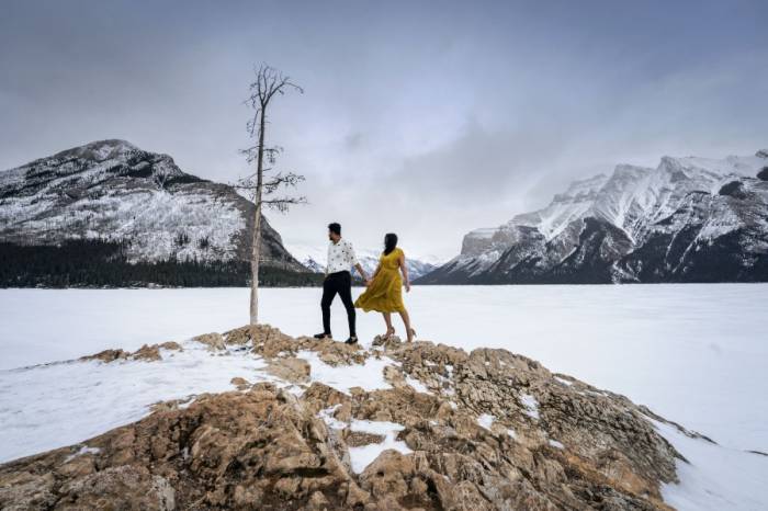 Engaged couple standing at the giant rock by frozen Minnewanka Lake. They look forward them and admire mountains.