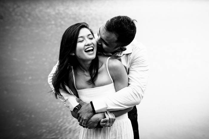 Man whisper a secret to his fiancee ear. She laughs. Black and white photography taken by best Banff Wedding Photographer.