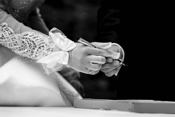 Black and white photograph of newlyweds hands. Bride passing a pen to the groom.