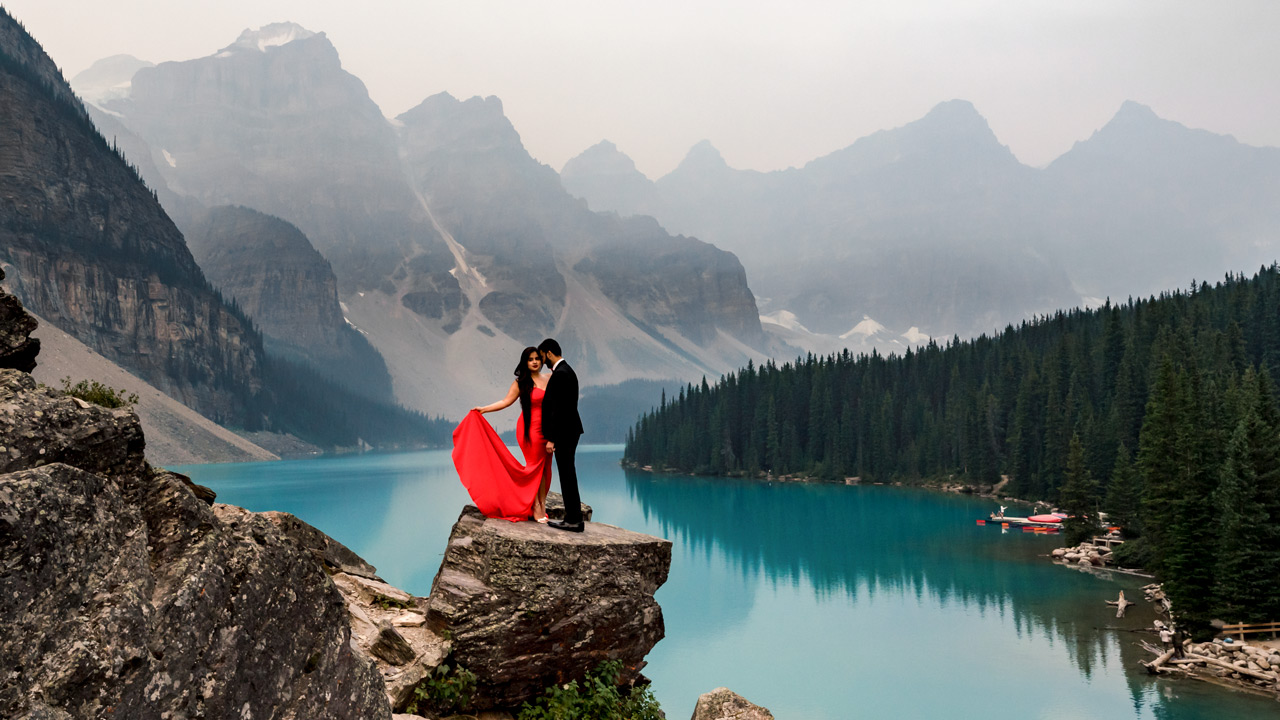 Moraine Lake Engagement Photography. Indian engaged couple standing on the rock. Lady is wearing a red, long dress and her fiancée is wearing a suite. | 4Eyes Photography