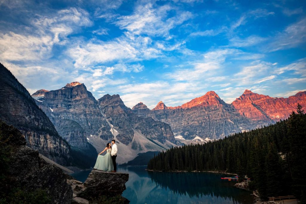 Bride and groom on the rock by Moraine Lake. Bride plays with her wedding dress while the groom look at her. Behind them tremendous Canadian Rockies. | 4Eyes Photography