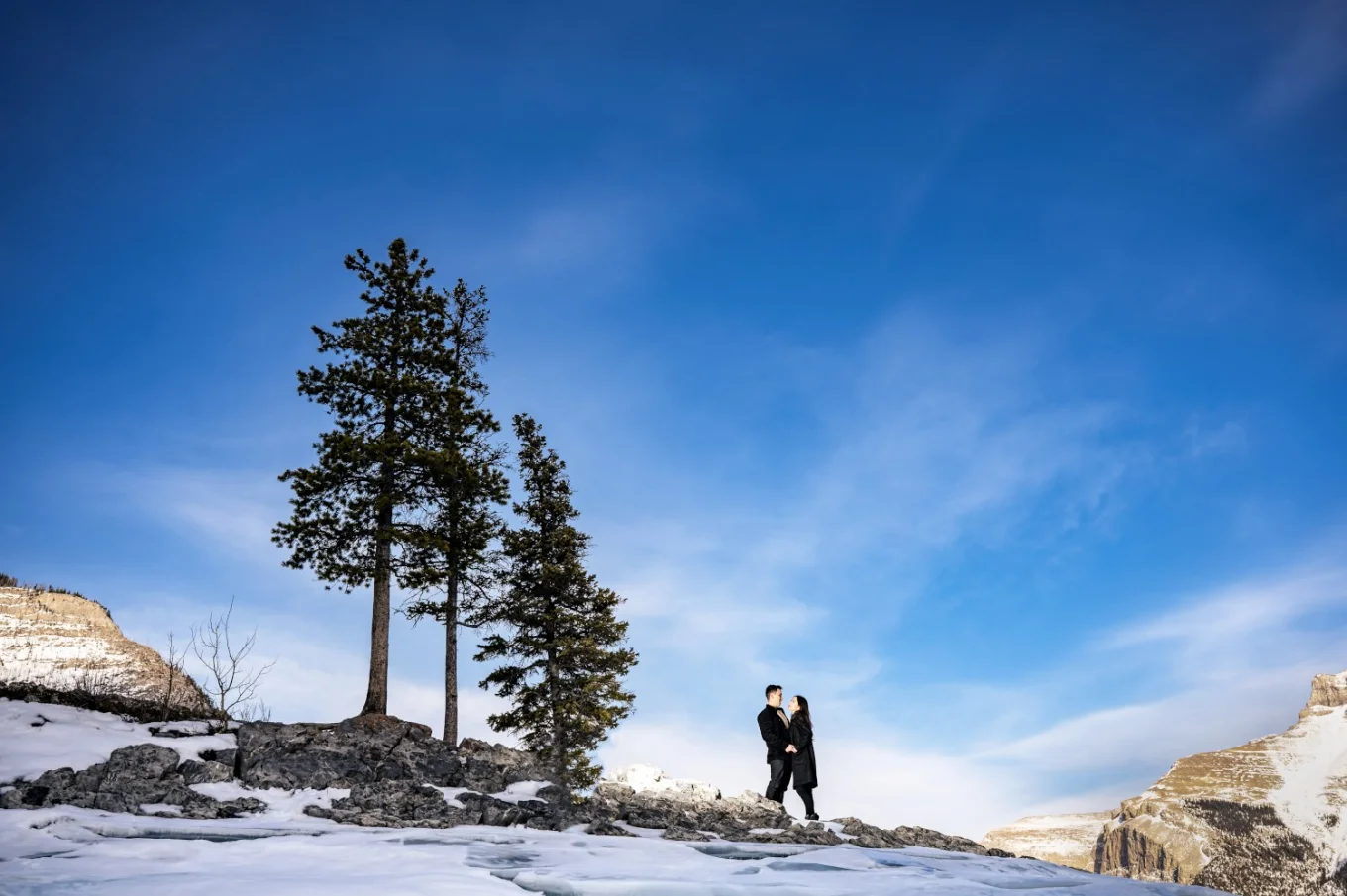 Tiny people in a hug up the hill. Next to them three trees and blue sky. Photograph taken in Banff by Calgary Wedding Photographer.