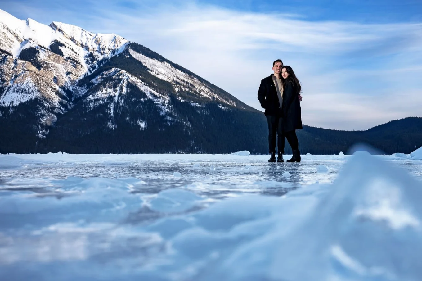 Young man and a woman in a hug at the frozen Minnewanka Lake. Behind them giant mountain.