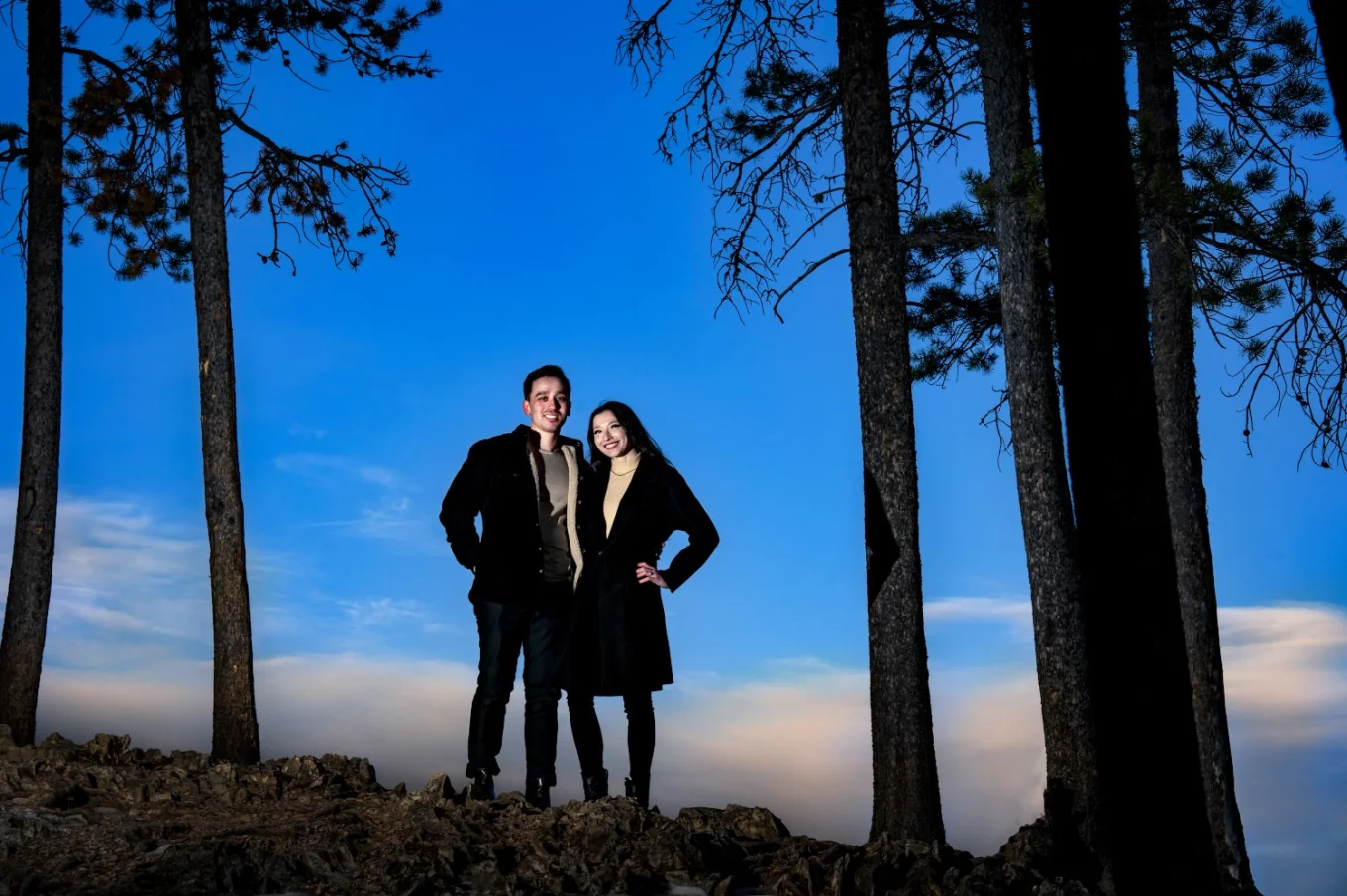 Handsome lovers posing for Calgary Wedding Photographer at Minnewanka Lake. They stand up the hill, between trees during sunset.