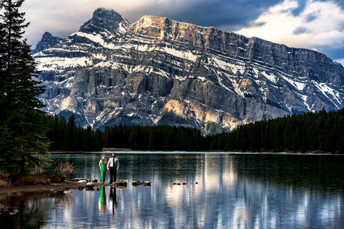 Woman in a green dress and her fiance hold their hands at the stunning Two Jack Lakes in Banff. In the background tremendous mountain.