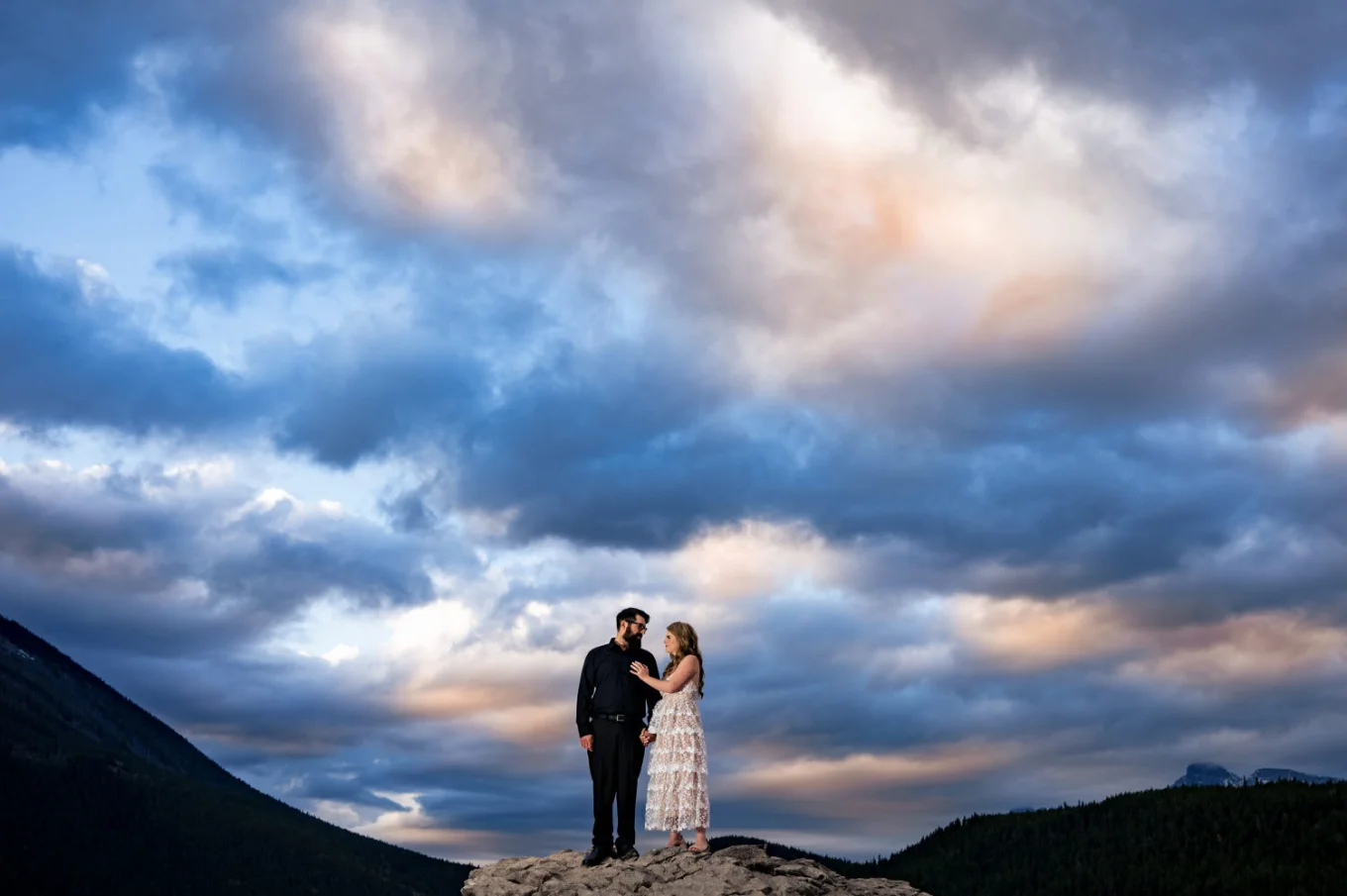 Woman in a white dress touches her fiance's chest and look deeply into his eyes. Above them dramatic sky.