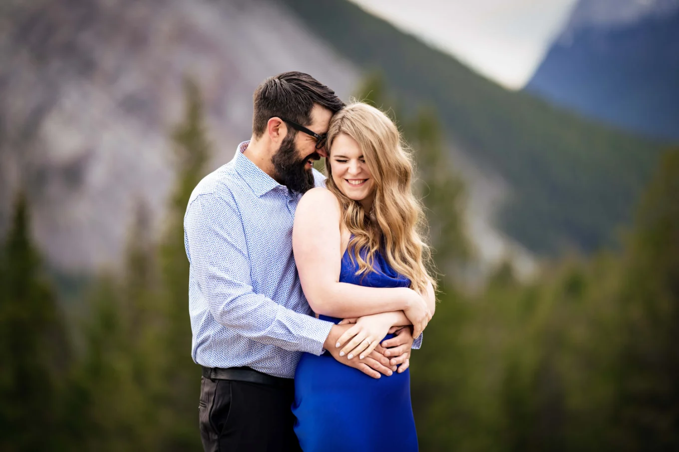 Man with long black beard whispers a secret into his fiancee's ear. They both laugh.