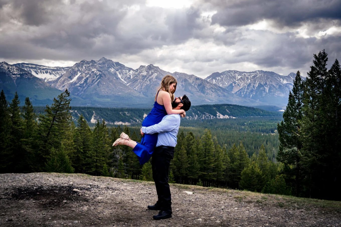 Gentleman lifts his fiancee. She wears a blue dress. They are in the mountains in Banff National Park.