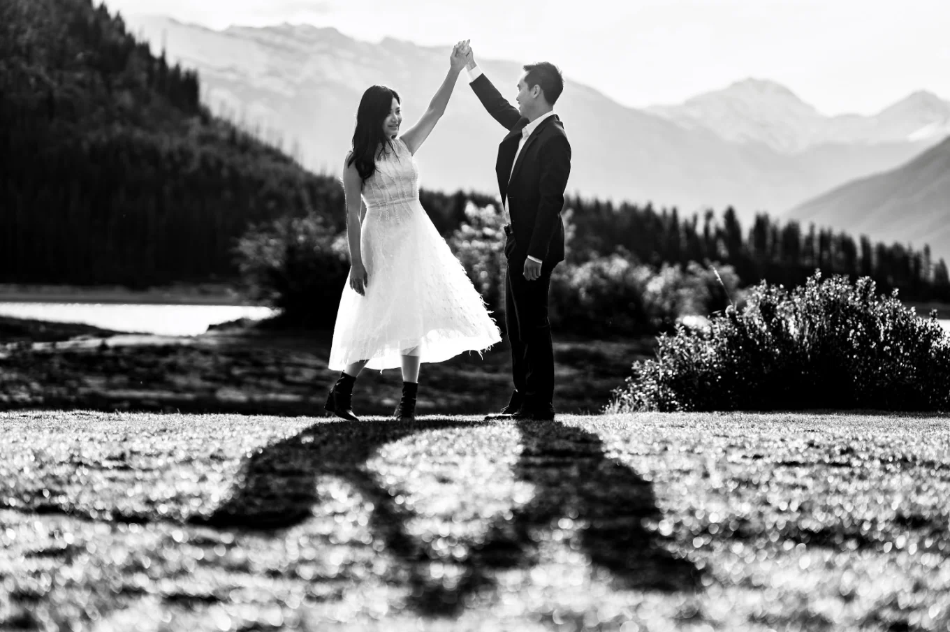 Creative photograph in black and white taken by Banff Wedding Photographer. Couple dance by the lake while they shadows are visible at the ground.