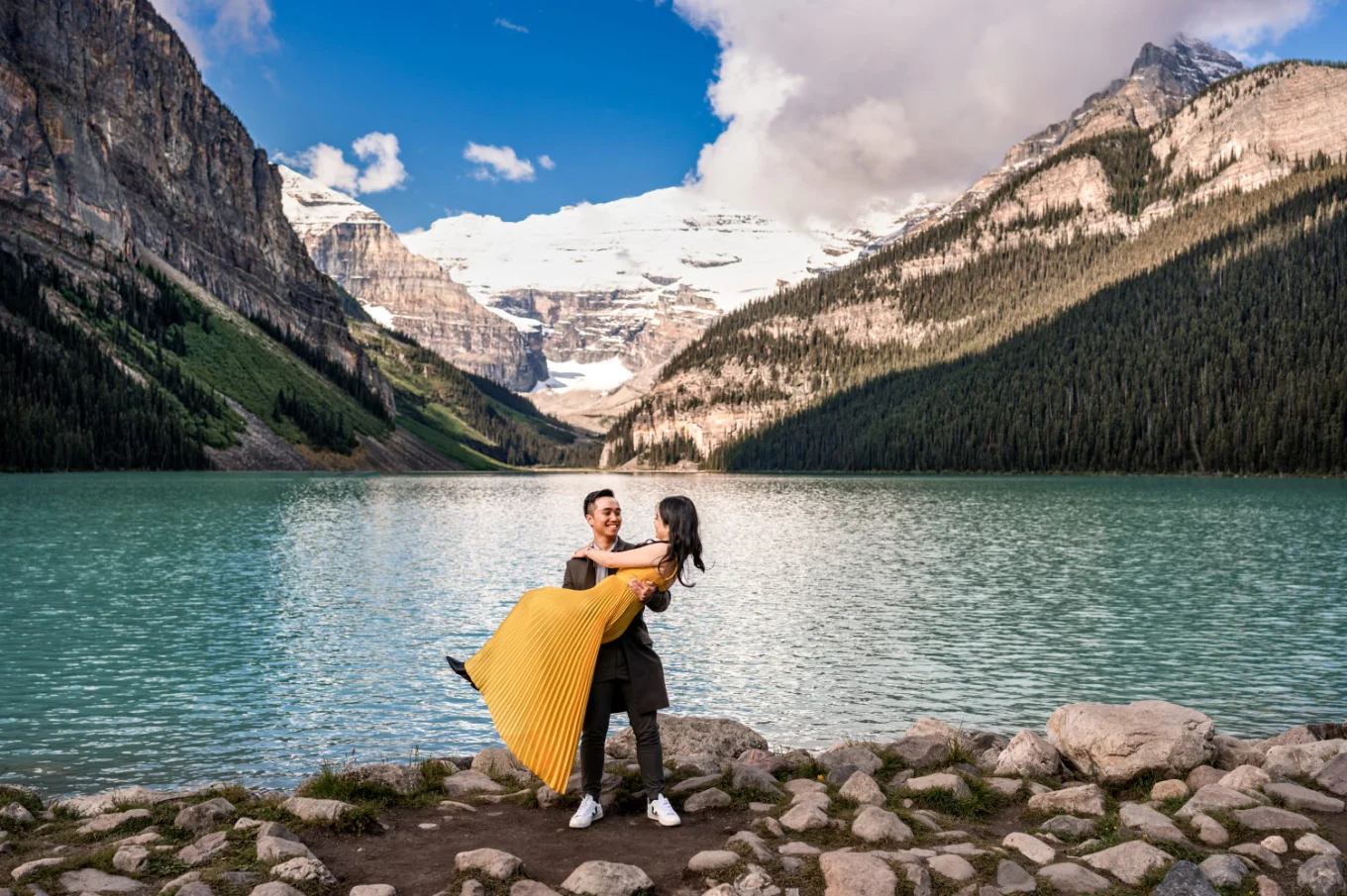 Man lifting his fiancee in a yellow dress at the stunning location of Lake Louise. Behind them lake and the Canadian Rockies.