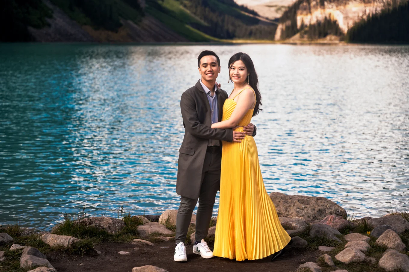 Engaged couple stand in ahead and look at the camera by the lake.
