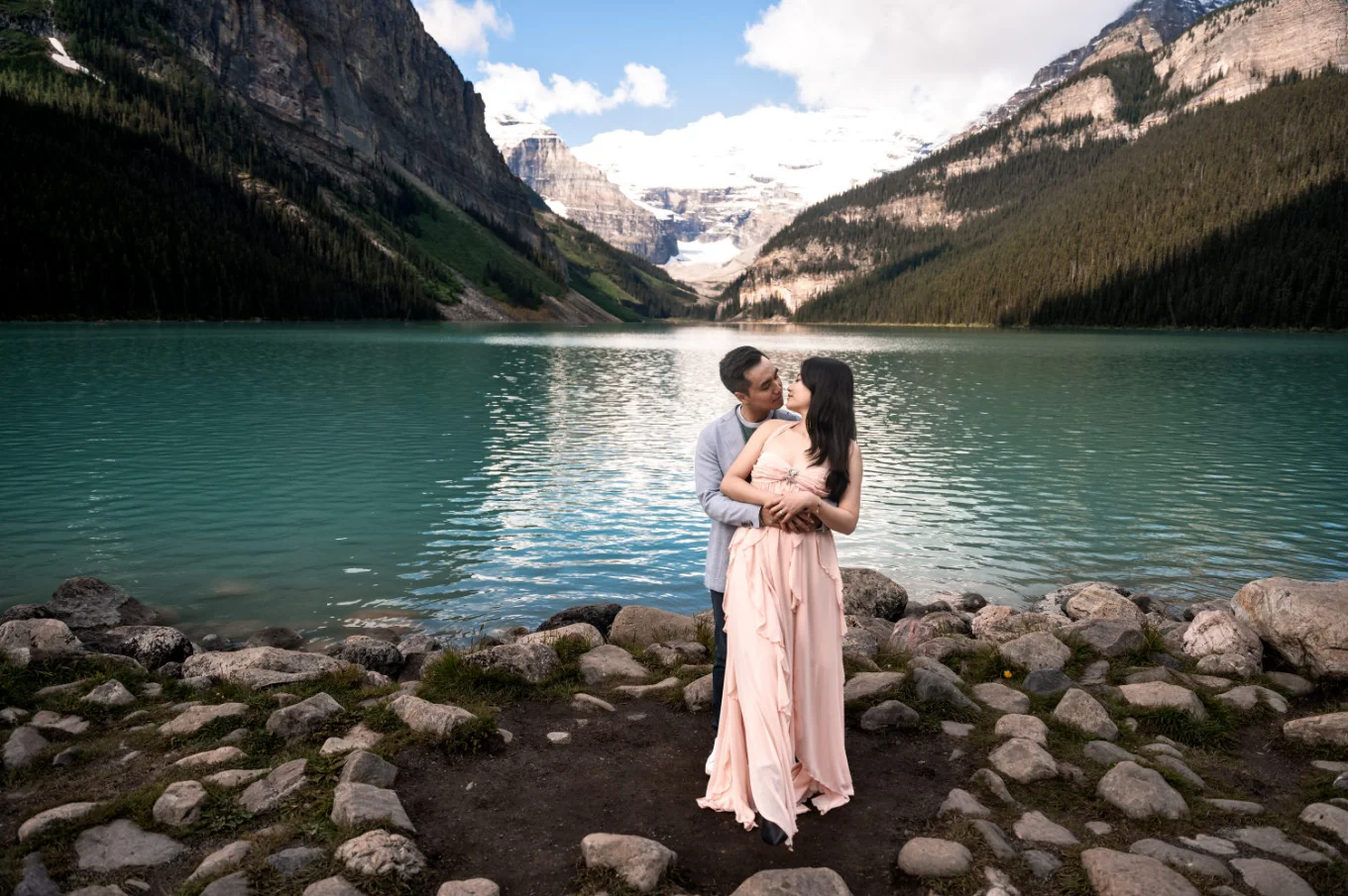 A man hug his fiancee from behind, they look at each other at the edge on Lake Louise.