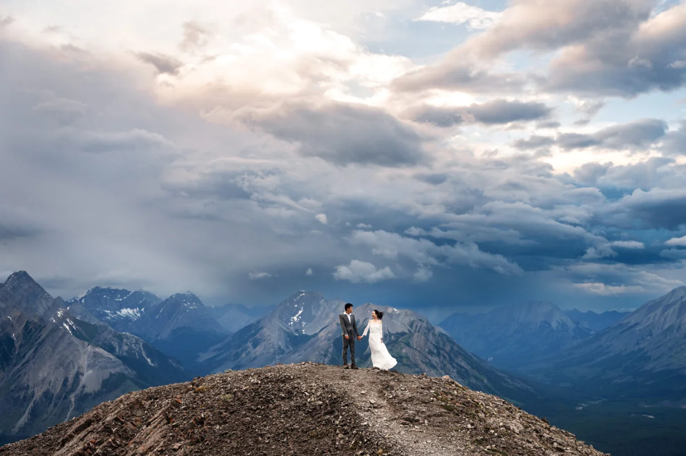 Tiny bride and groom at the ridge of the mountain siding sunset with the heavy clouds above them.