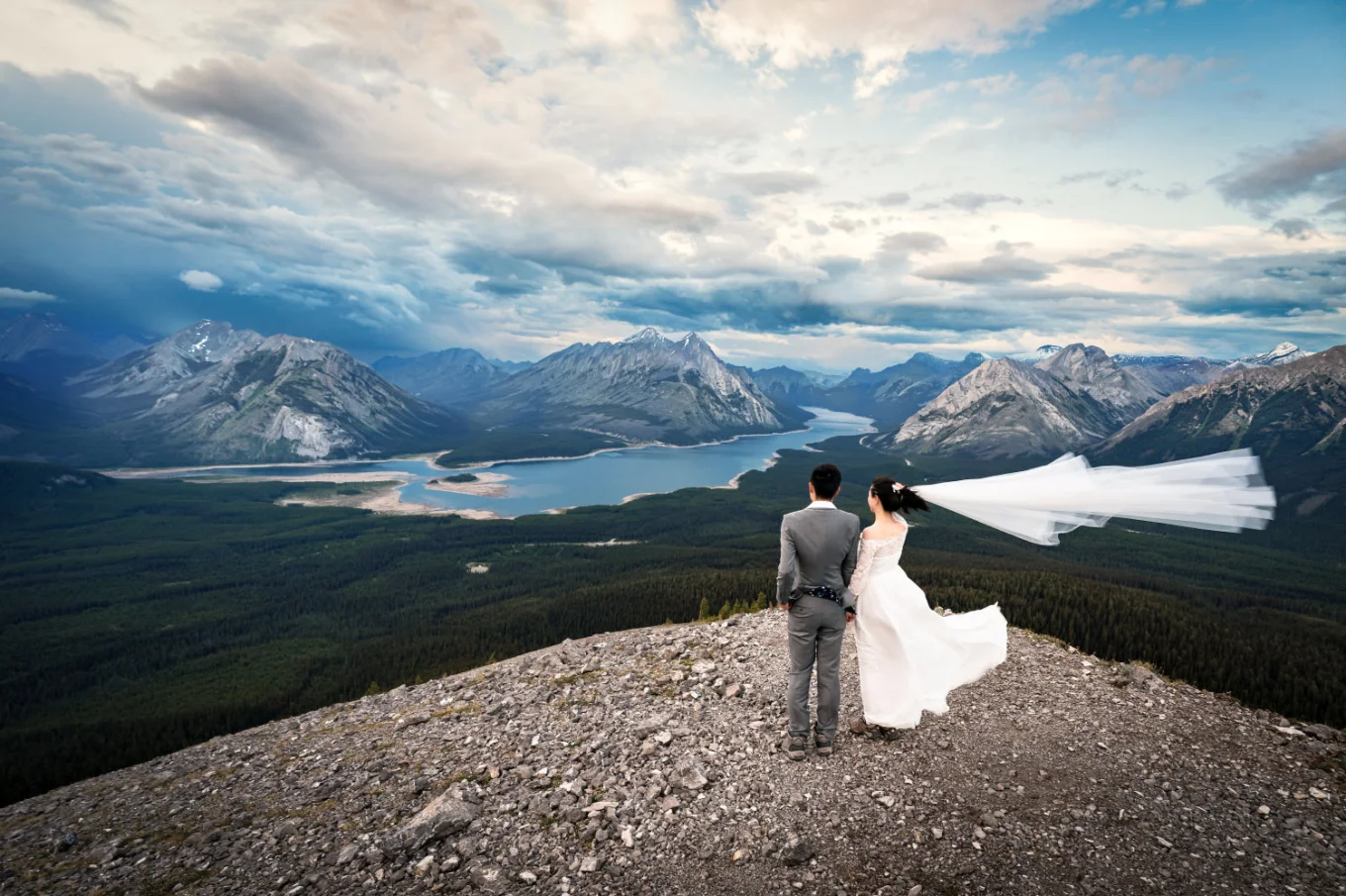 Bride and groom adore the stunning view of the Canadian Rockies and a blue lake from the peak.