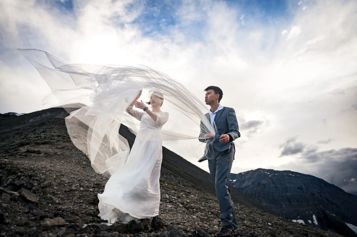 The groom helps the bride to put back her veil during windy day in the mountains.