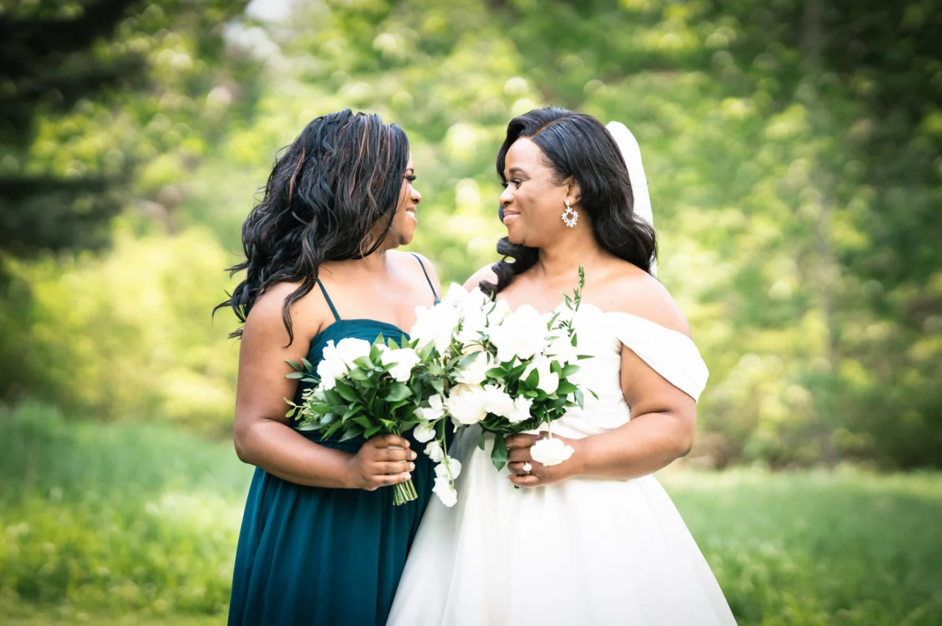 Portrait on the bride and maid of honour looking at each other. They hold white flowers in their hands.