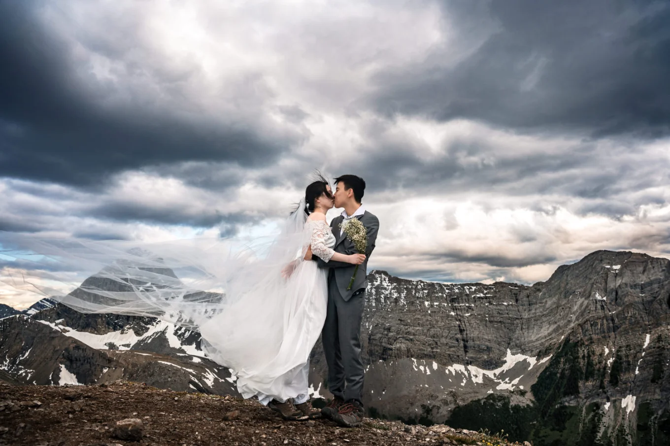 Bride in a long white dress and the groom share a kiss in Banff National Park. Above them heavy clouds during the sunset.