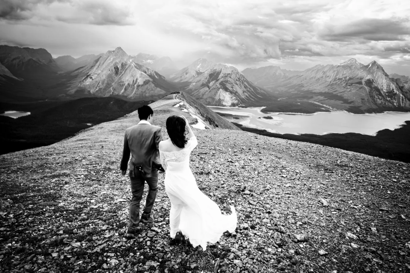 Newlyweds walk at the ridge of Tent Ridge with majestic view of the Rocky Mountains and the lake.