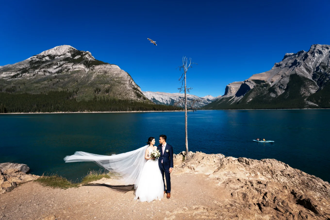 Stunning wedding portrait at Minnewanka Lake taken by Banff Wedding Photographer. Newlyweds standing next to each other a look at their eyes. Behind them Blue lake and Rocky Mountains.
