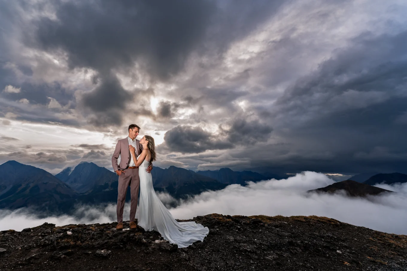 Bride hug the groom at the summit of the mountain in Banff. On the background huge Rocky Mountains and heavy clouds.