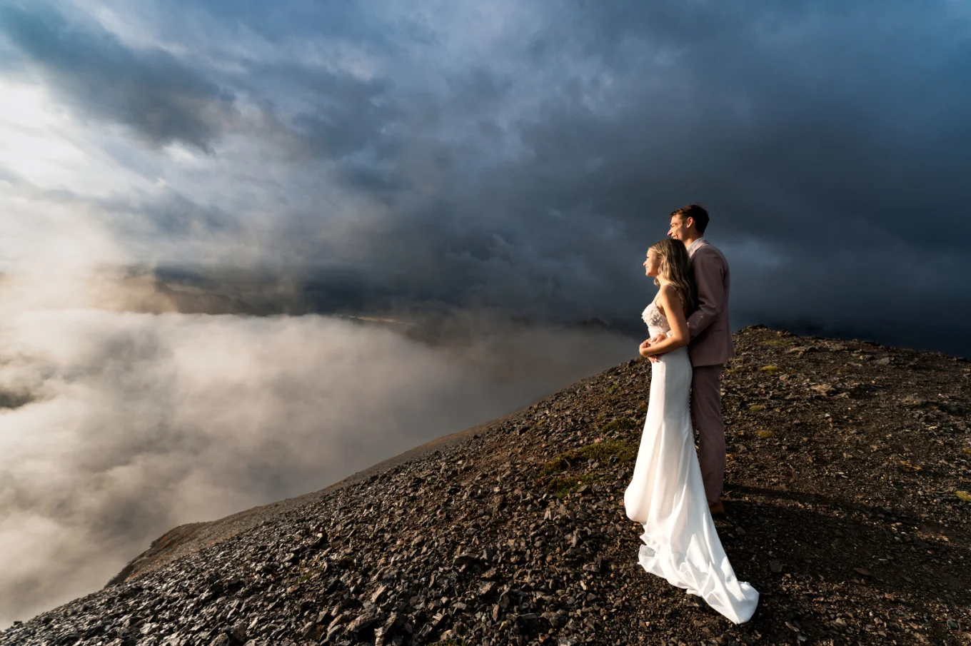 A blissful wedding couple stands atop a mountain, surrounded by ethereal clouds, embracing their love amidst breathtaking scenery in Banff.